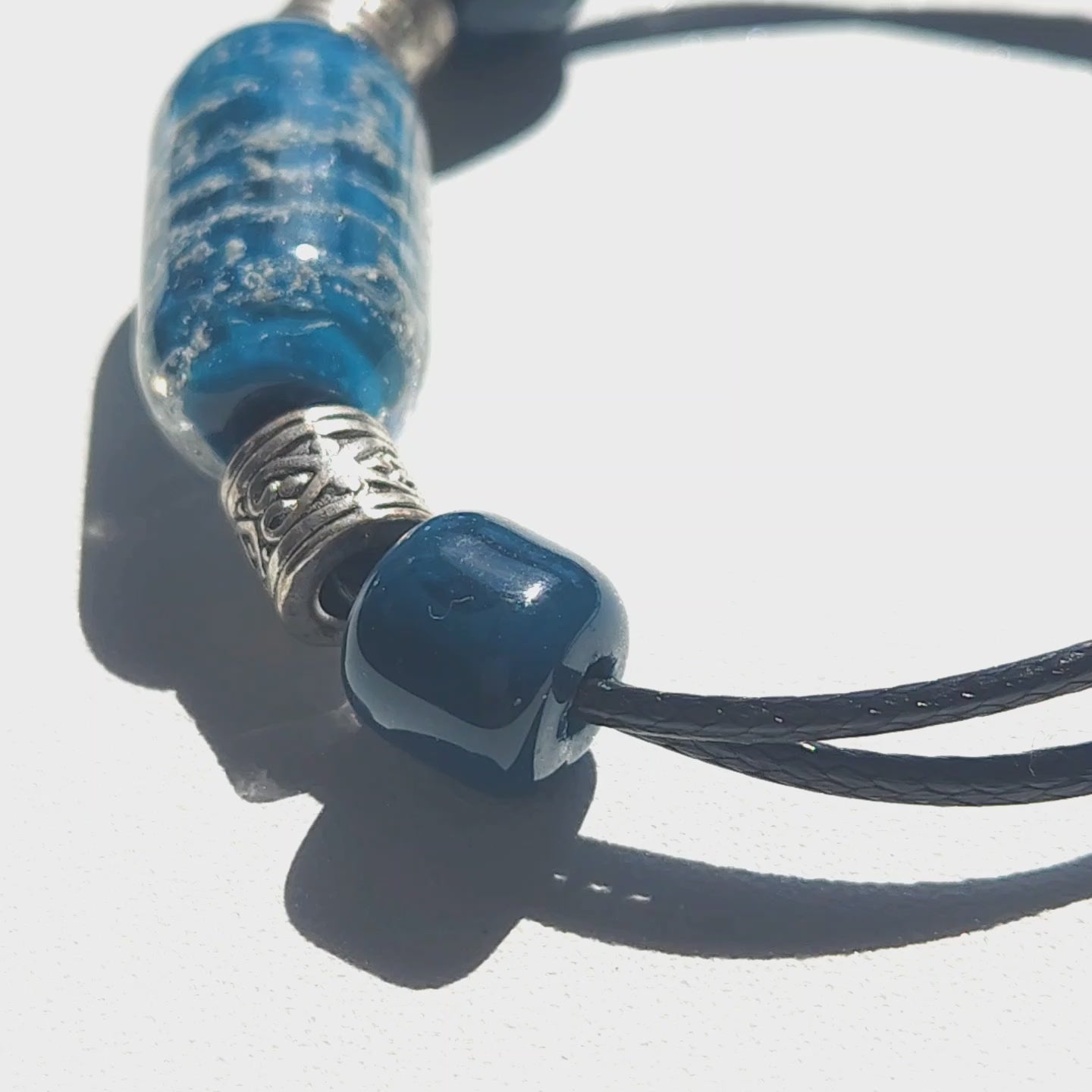 Close-up of blue Remember Me Urn Bracelet with ashes encased in cremation bead, featuring silver accents and black cord. Cremation jewelry for memorial use.