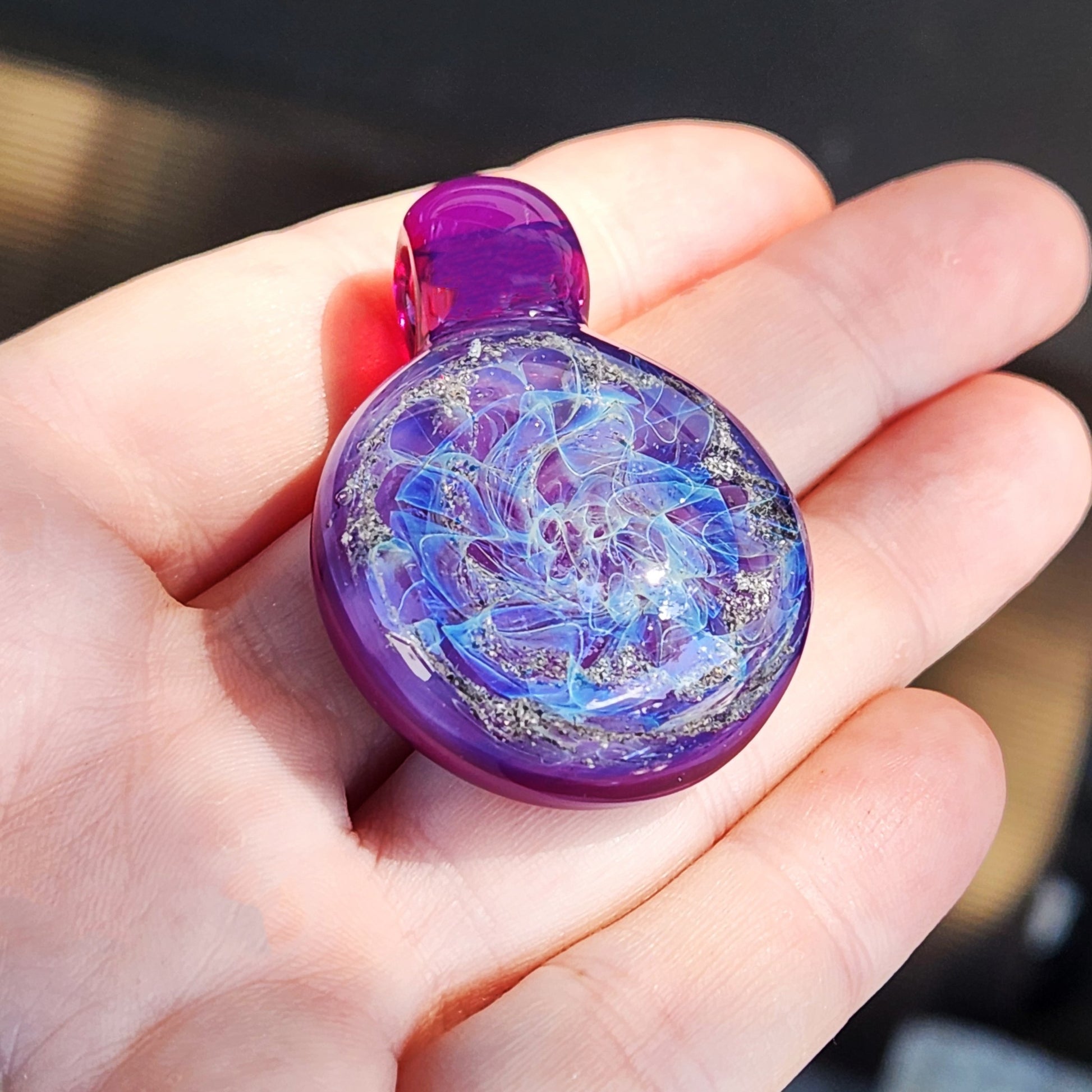 A purple glass memorial pendant with a swirling, galaxy-like design. The cremation pendant features shades of purple, blue, and ashes, with a rounded shape and a dark purple stem. A black cord is attached, completing the urn necklace. The glass pendant is a unique piece of memorial jewelry.