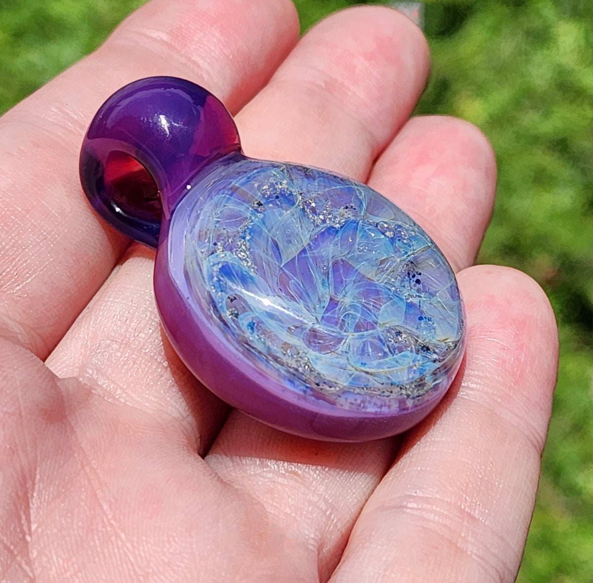 A purple glass memorial pendant with a swirling, galaxy-like design. The cremation pendant features shades of purple, blue, and ashes, with a rounded shape and a dark purple stem. A black cord is attached, completing the urn necklace. The glass pendant is a unique piece of memorial jewelry.