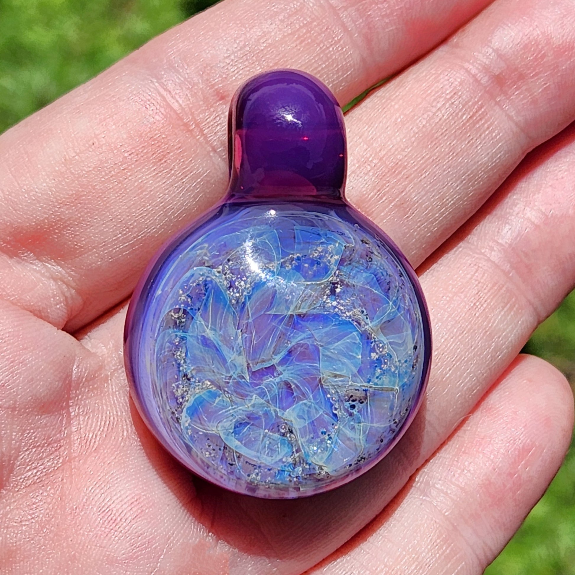 A purple glass memorial pendant with a swirling, galaxy-like design. The cremation pendant features shades of purple, blue, and ashes, with a rounded shape and a dark purple stem. A black cord is attached, completing the urn necklace. The glass pendant is a unique piece of memorial jewelry.