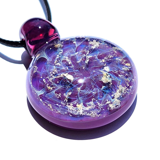 A purple glass memorial pendant with a swirling, galaxy-like design. The cremation pendant features shades of purple, blue, and ashes, with a rounded shape and a dark purple stem. A black cord is attached, completing the urn necklace. The glass pendant is a unique piece of memorial jewelry.
